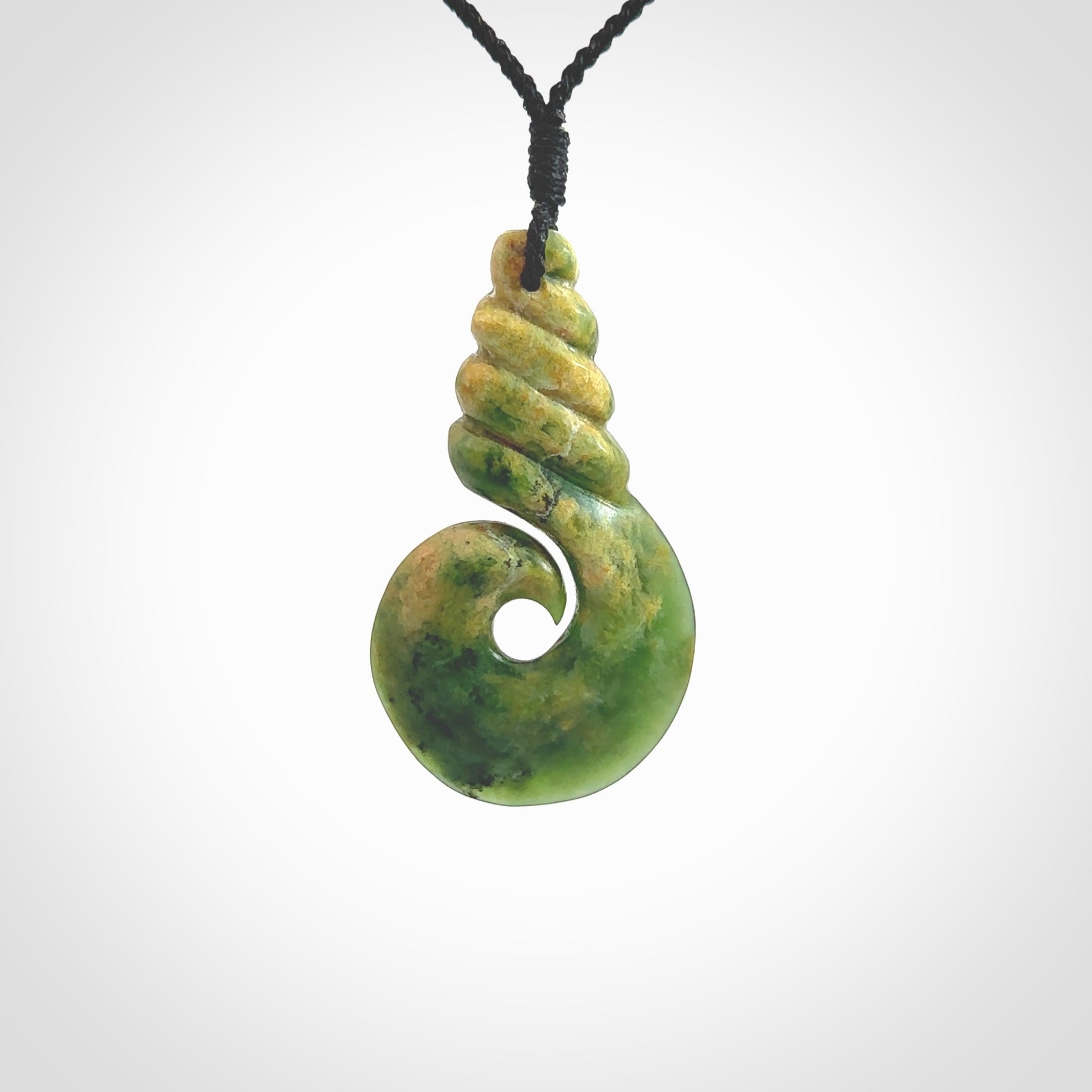 This picture shows a hook shaped pendant, hand carved from New Zealand flower jade. The top of the hook, the shaft, is a twist shape. We will provide this with an adjustable plaited cord. The cord is a adjustable and is a black colour. Twist and koru designs are also displayed in this stunning New Zealand Marsden Flower Jade pendant. Hand made by Ana Krakosky.