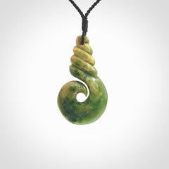 This picture shows a hook shaped pendant, hand carved from New Zealand flower jade. The top of the hook, the shaft, is a twist shape. We will provide this with an adjustable plaited cord. The cord is a adjustable and is a black colour. Twist and koru designs are also displayed in this stunning New Zealand Marsden Flower Jade pendant. Hand made by Ana Krakosky.