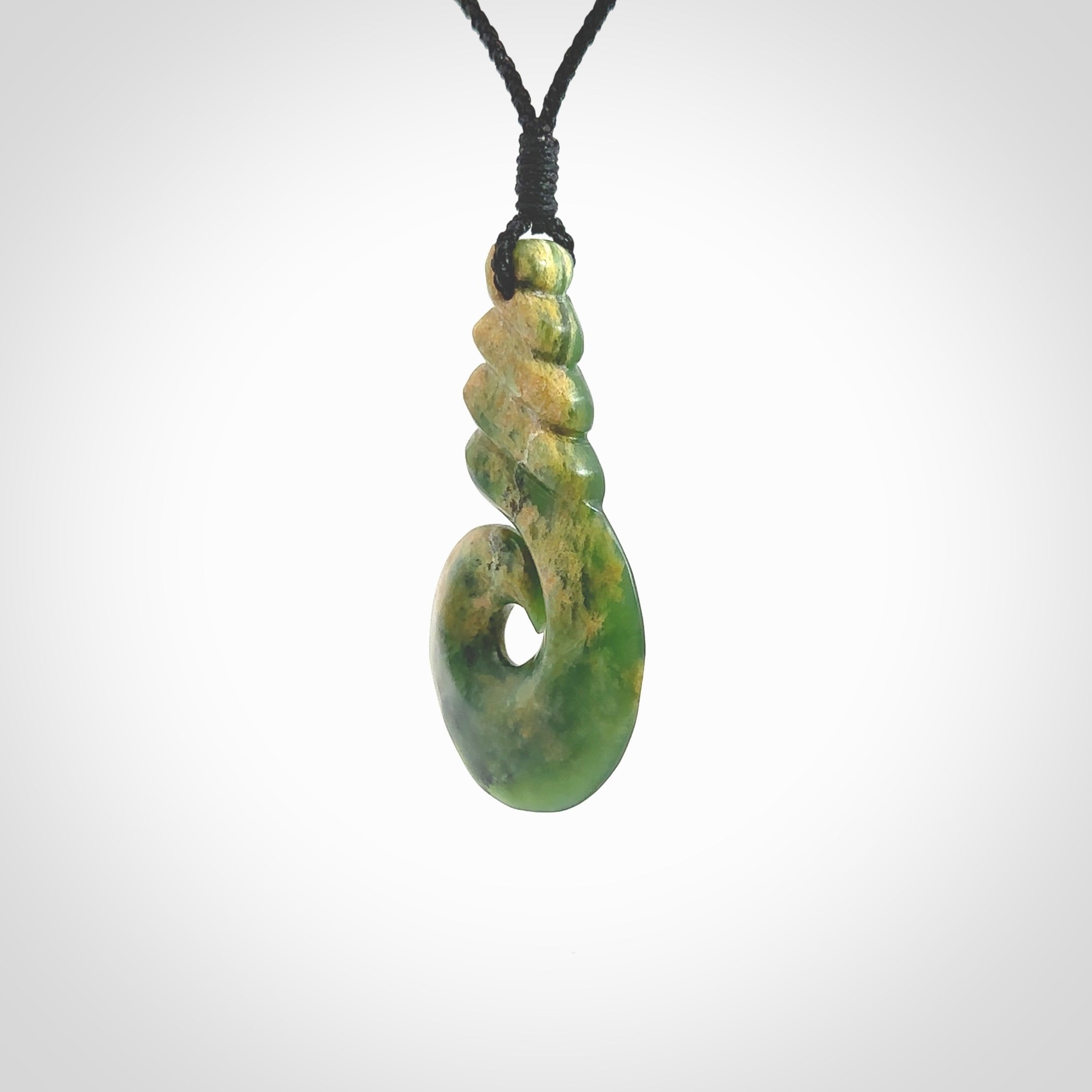 This picture shows a hook shaped pendant, hand carved from New Zealand flower jade. The top of the hook, the shaft, is a twist shape. We will provide this with an adjustable plaited cord. The cord is a adjustable and is a black colour. Twist and koru designs are also displayed in this stunning New Zealand Marsden Flower Jade pendant. Hand made by Ana Krakosky.