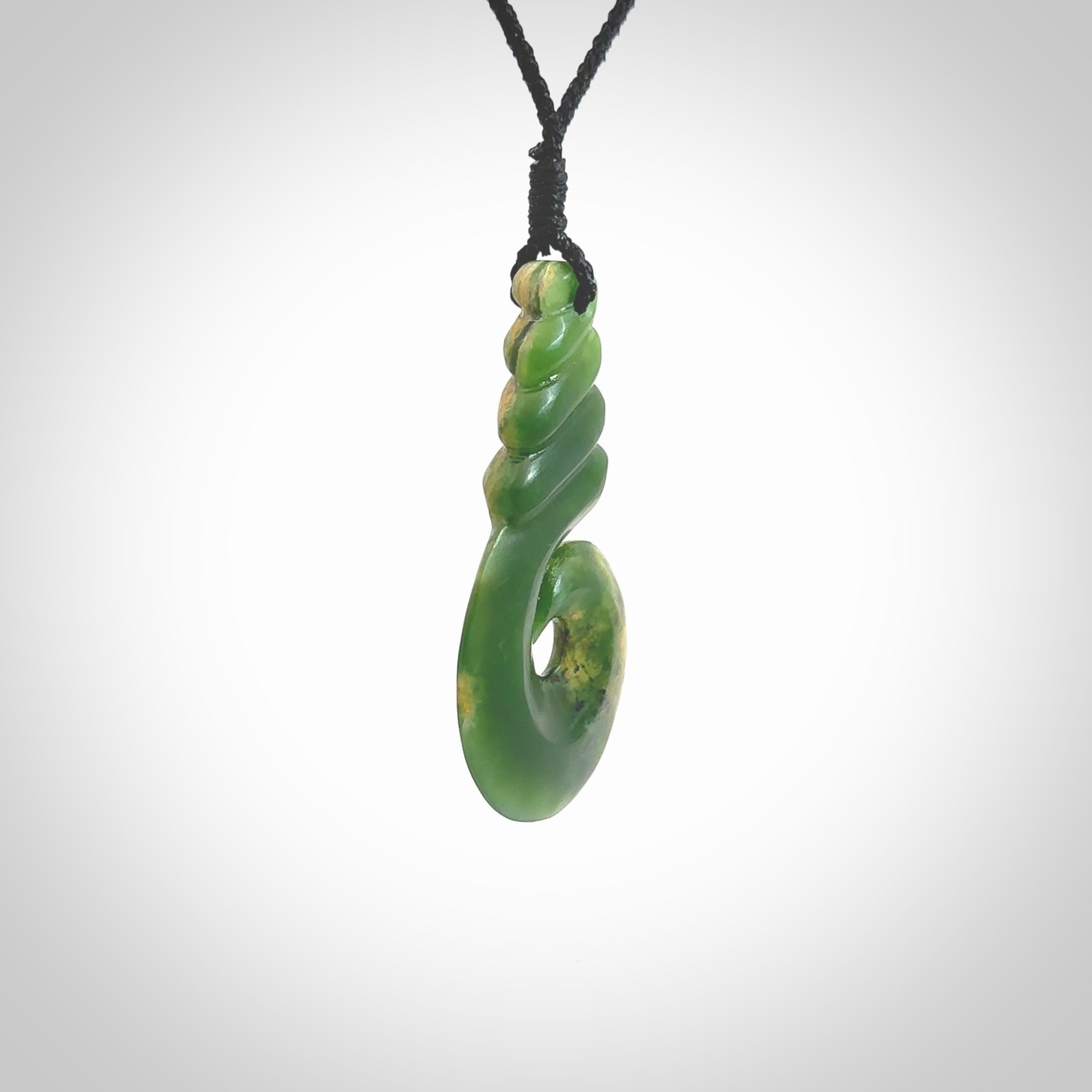 This picture shows a hook shaped pendant, hand carved from New Zealand flower jade. The top of the hook, the shaft, is a twist shape. We will provide this with an adjustable plaited cord. The cord is a adjustable and is a black colour. Twist and koru designs are also displayed in this stunning New Zealand Marsden Flower Jade pendant. Hand made by Ana Krakosky.