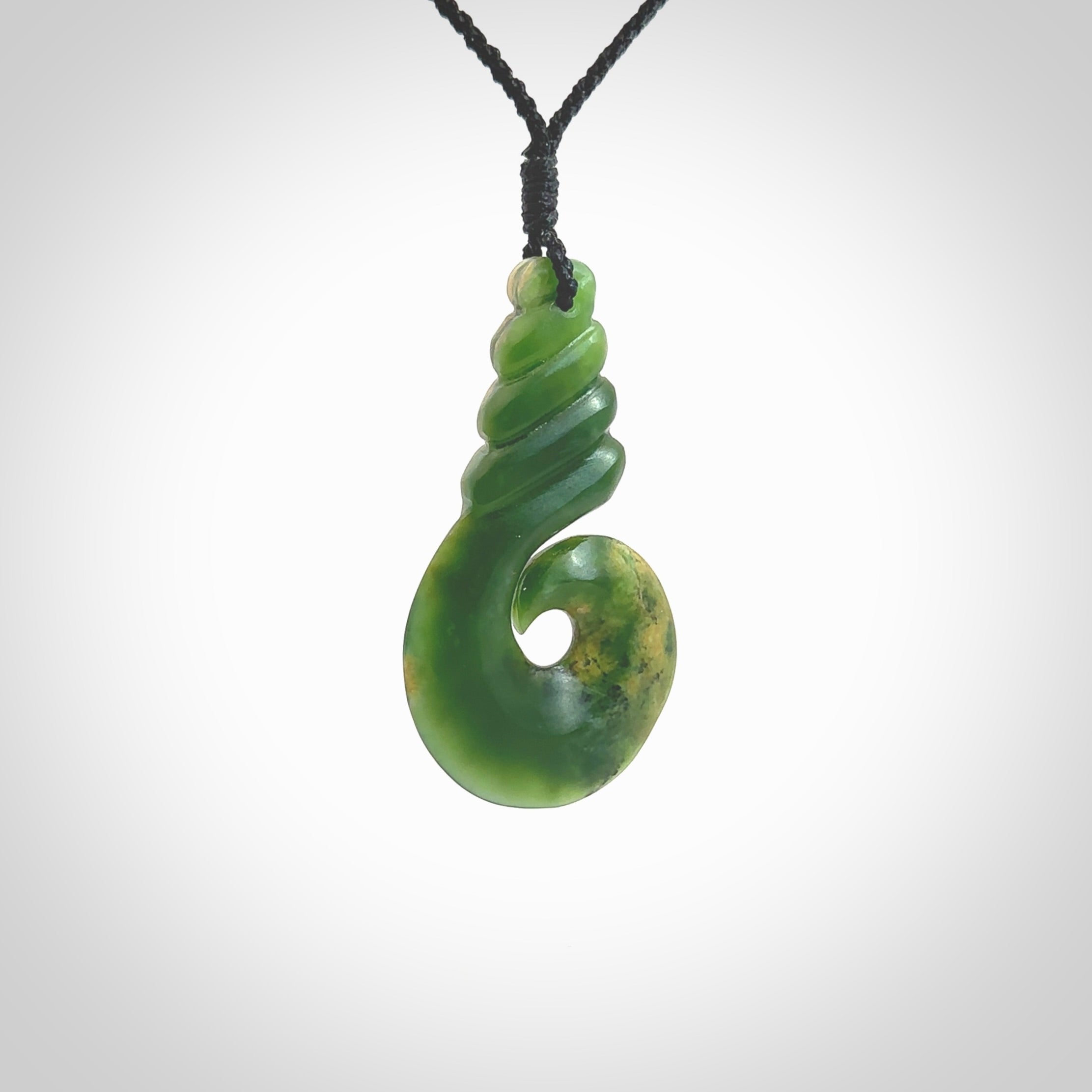 This picture shows a hook shaped pendant, hand carved from New Zealand flower jade. The top of the hook, the shaft, is a twist shape. We will provide this with an adjustable plaited cord. The cord is a adjustable and is a black colour. Twist and koru designs are also displayed in this stunning New Zealand Marsden Flower Jade pendant. Hand made by Ana Krakosky.
