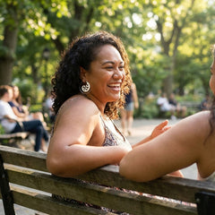 Two women sitting on a park bench laughing together with trees in the background showcasing bone earring