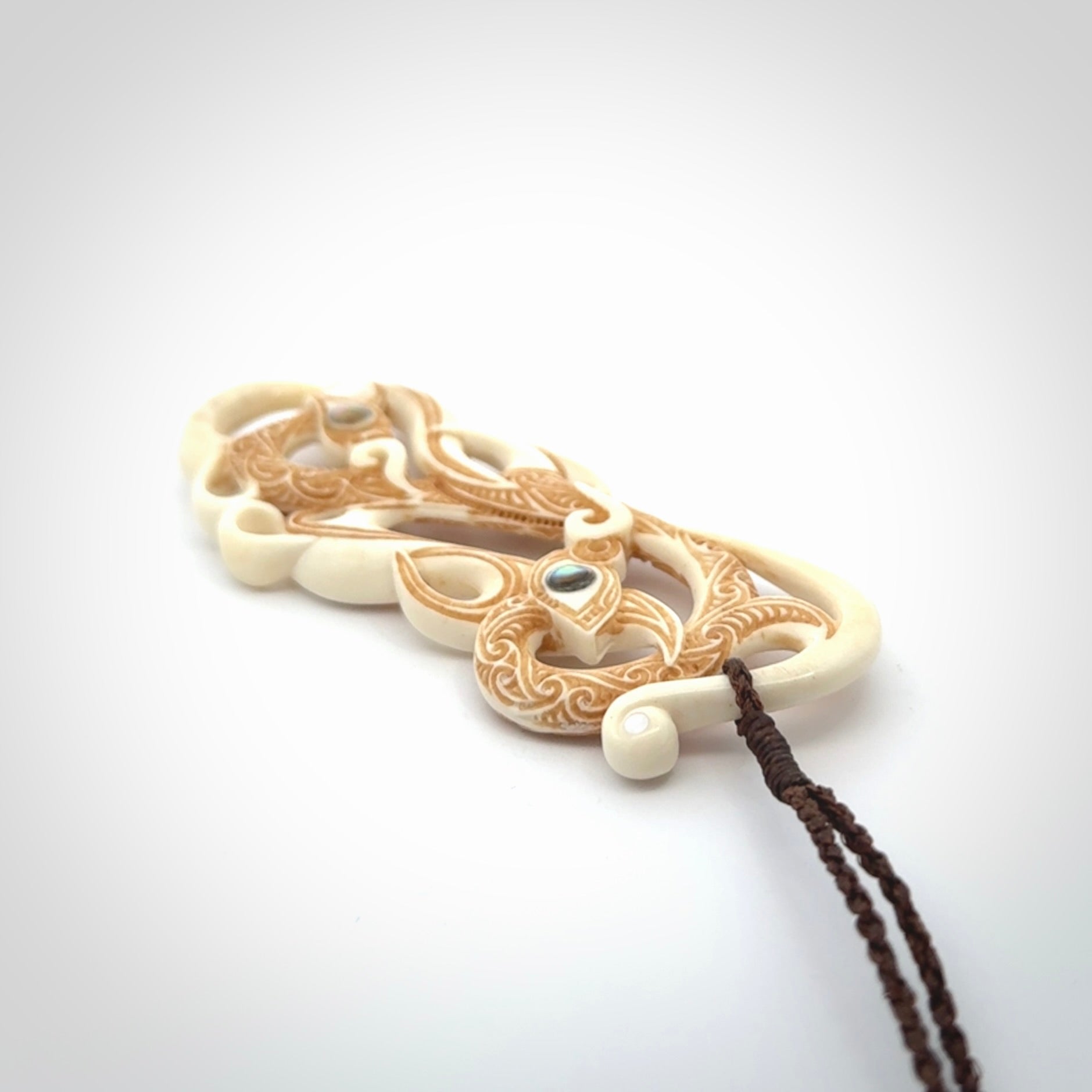 This picture shows a carved double manaia in bone. The artist has carved traditional decorative Koru designs into the face of the body and these run up the sides of the manaia head. These have specific meanings. It is provided with a hand-plaited brown cord that is length adjustable.