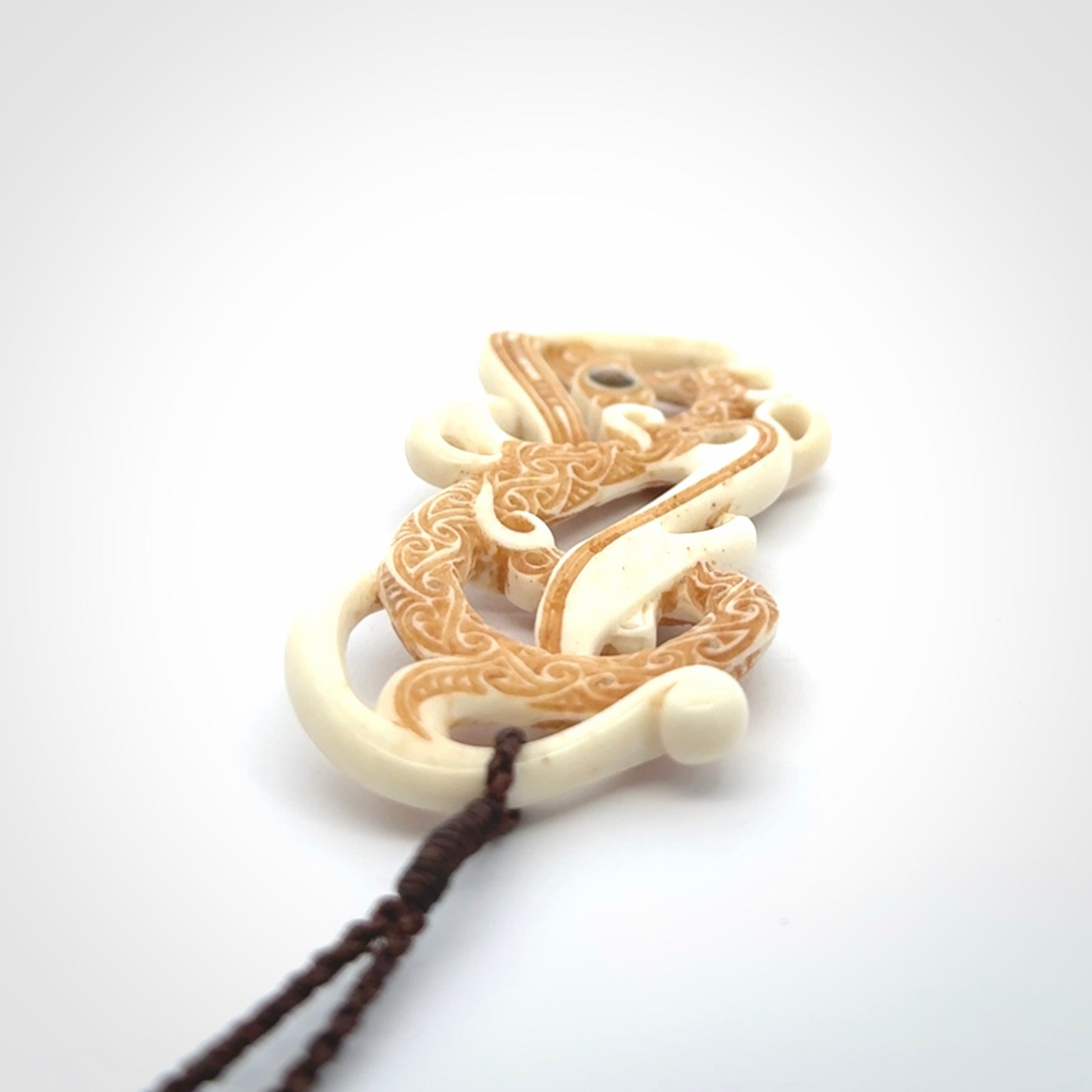 This picture shows a carved double manaia in bone. The artist has carved traditional decorative Koru designs into the face of the body and these run up the sides of the manaia head. These have specific meanings. It is provided with a hand-plaited brown cord that is length adjustable.