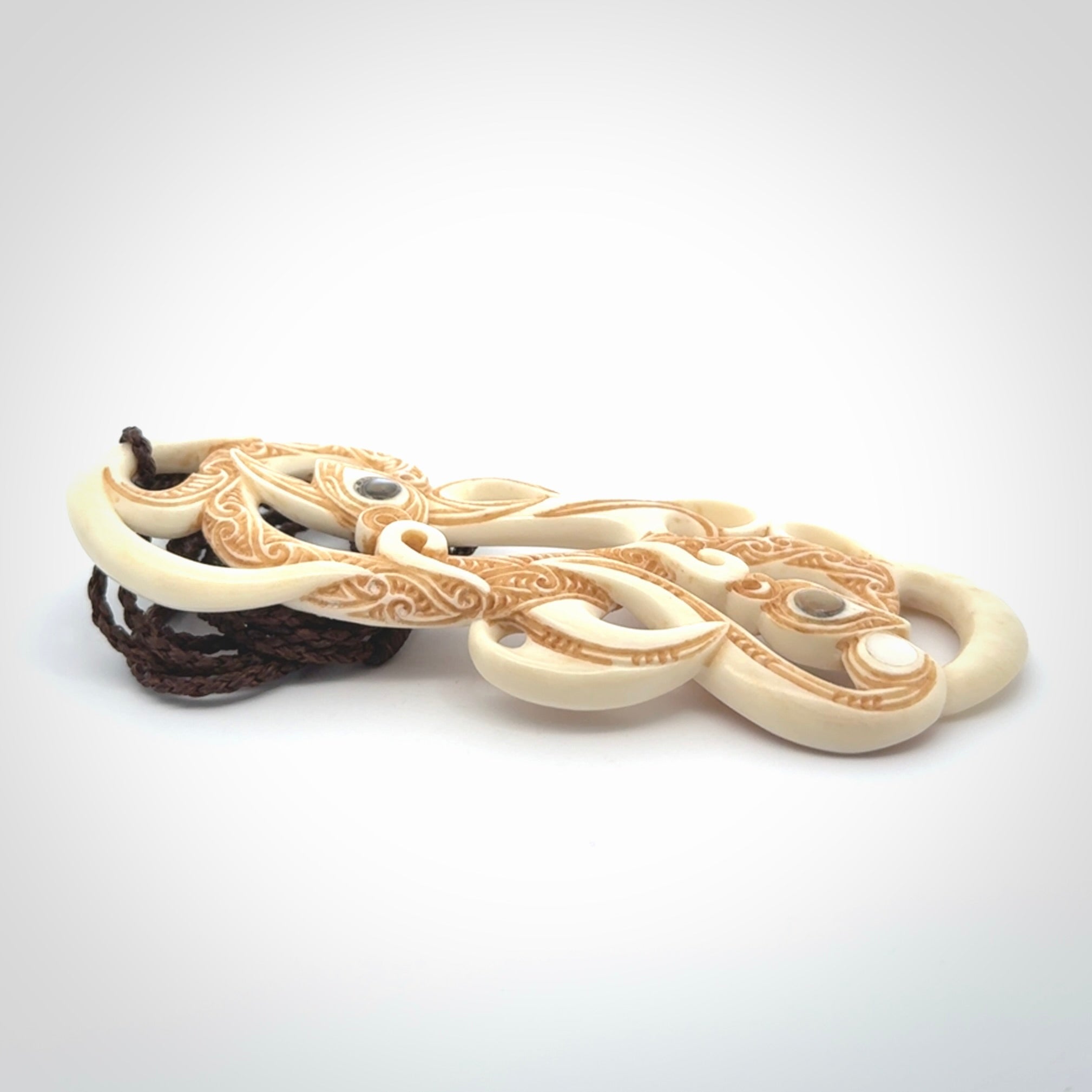 This picture shows a carved double manaia in bone. The artist has carved traditional decorative Koru designs into the face of the body and these run up the sides of the manaia head. These have specific meanings. It is provided with a hand-plaited brown cord that is length adjustable.