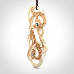 This picture shows a carved double manaia in bone. The artist has carved traditional decorative Koru designs into the face of the body and these run up the sides of the manaia head. These have specific meanings. It is provided with a hand-plaited brown cord that is length adjustable.