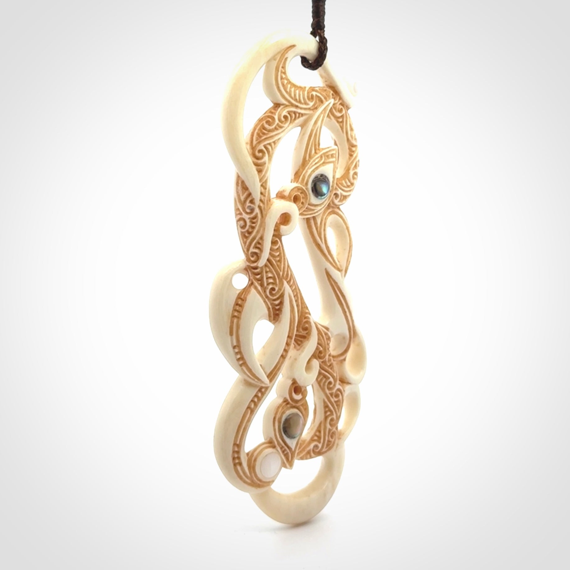 This picture shows a carved double manaia in bone. The artist has carved traditional decorative Koru designs into the face of the body and these run up the sides of the manaia head. These have specific meanings. It is provided with a hand-plaited brown cord that is length adjustable.