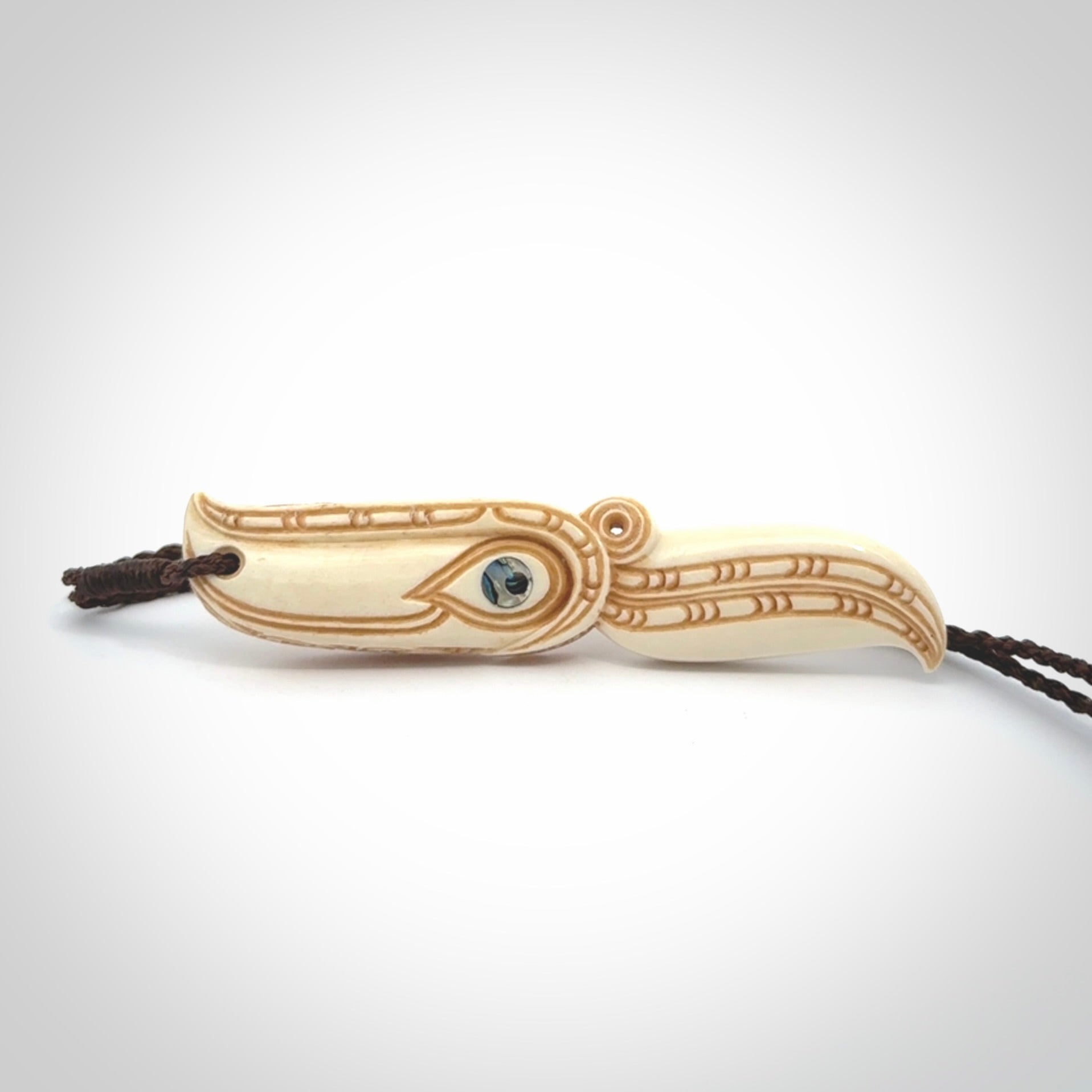 This picture shows a carved hawk in bone. It is a drop shaped pendant. The artist has carved traditional decorative kowhaiwhai designs into the body and these run up the sides of the hawk. These have specific meanings. It is provided with a hand-plaited brown cord that is length adjustable. Shipping is included and we ship worldwide.