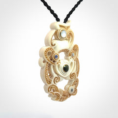 Hand carved bone manaia pendant with Jade, Mother of Pearl, Paua shell and Brass inlay. This is a complex carving by New Zealand carver and artist, Andrew Doughty. It is an intricate representation of a manaia carved to represent protection. It can be worn but is also a quality that warrants display.