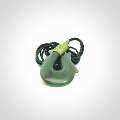 This photo shows a greenstone hook, or matau, pendant. It is a beautiful green British Columbia jade stone. The plaited cord is a green colour. It is plaited and the length can be adjusted.