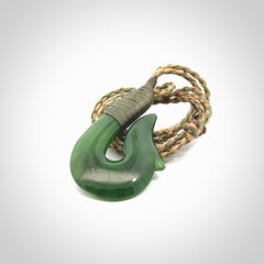 This photo shows a greenstone hook, or matau, pendant. It is a beautiful green British Columbia jade stone. The plaited cord is a green and tan colour. It is plaited and the length can be adjusted.