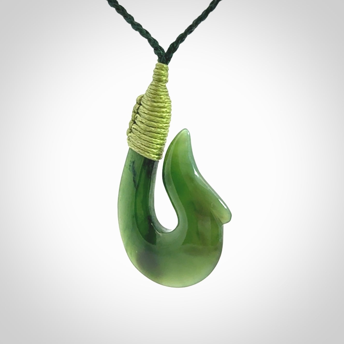 This photo shows a greenstone hook, or matau, pendant. It is a beautiful green British Columbia jade stone. The plaited cord is a green colour. It is plaited and the length can be adjusted.