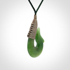 This photo shows a greenstone hook, or matau, pendant. It is a beautiful green British Columbia jade stone. The plaited cord is a green colour. It is plaited and the length can be adjusted.