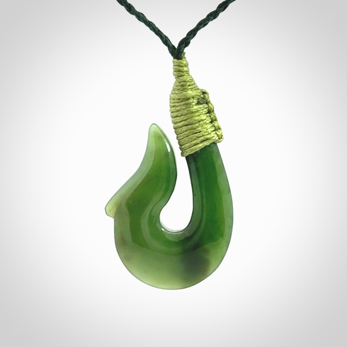 This photo shows a greenstone hook, or matau, pendant. It is a beautiful green British Columbia jade stone. The plaited cord is a green colour. It is plaited and the length can be adjusted.