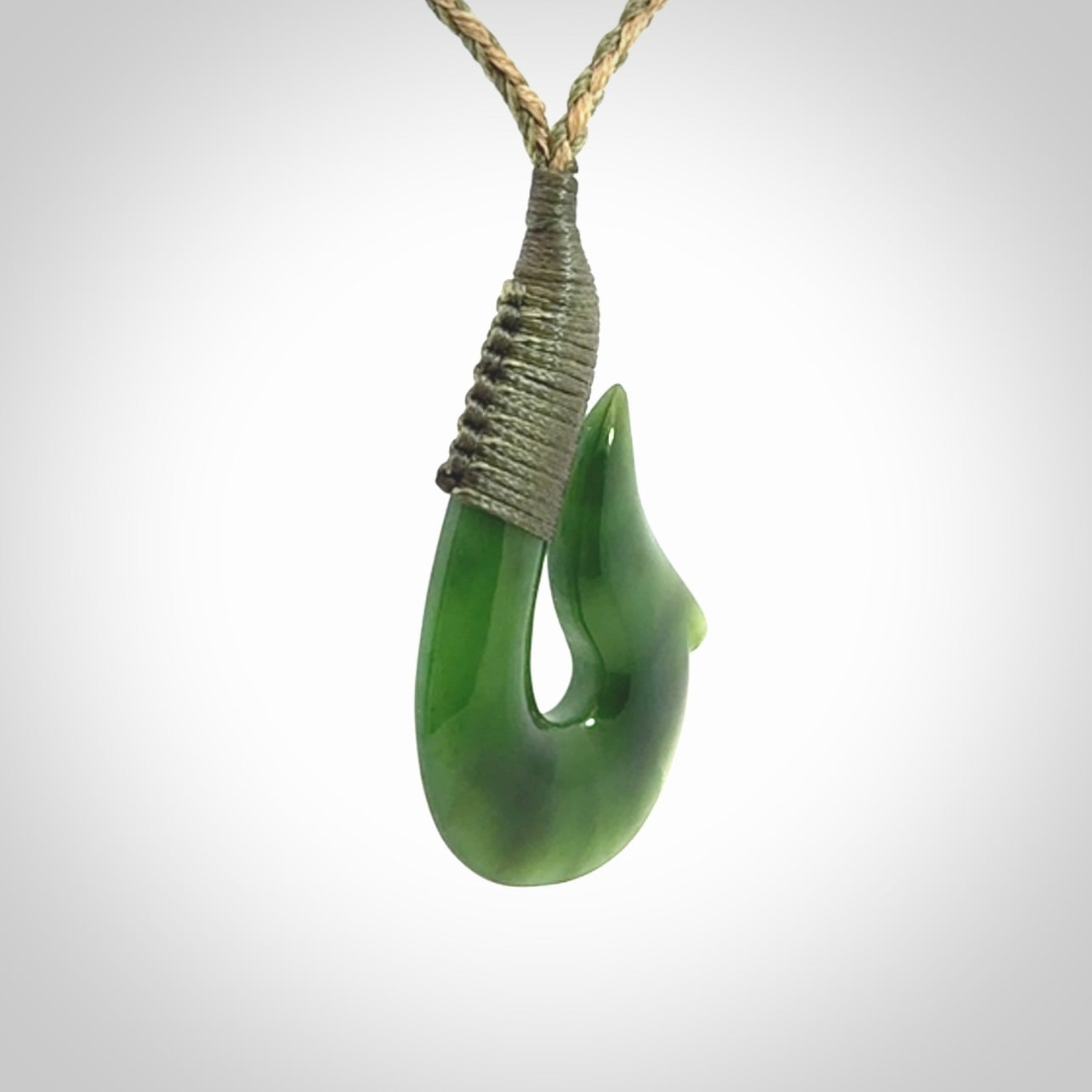 This photo shows a greenstone hook, or matau, pendant. It is a beautiful green British Columbia jade stone. The plaited cord is a green and tan colour. It is plaited and the length can be adjusted.