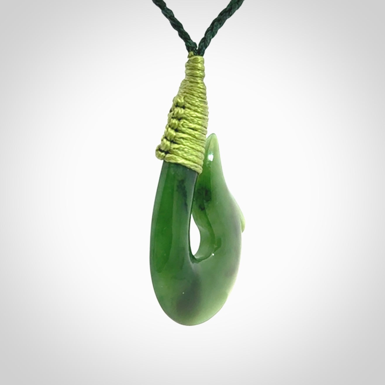 This photo shows a greenstone hook, or matau, pendant. It is a beautiful green British Columbia jade stone. The plaited cord is a green colour. It is plaited and the length can be adjusted.