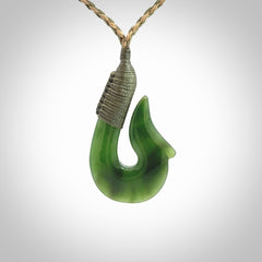 This photo shows a greenstone hook, or matau, pendant. It is a beautiful green British Columbia jade stone. The plaited cord is a green and tan colour. It is plaited and the length can be adjusted.