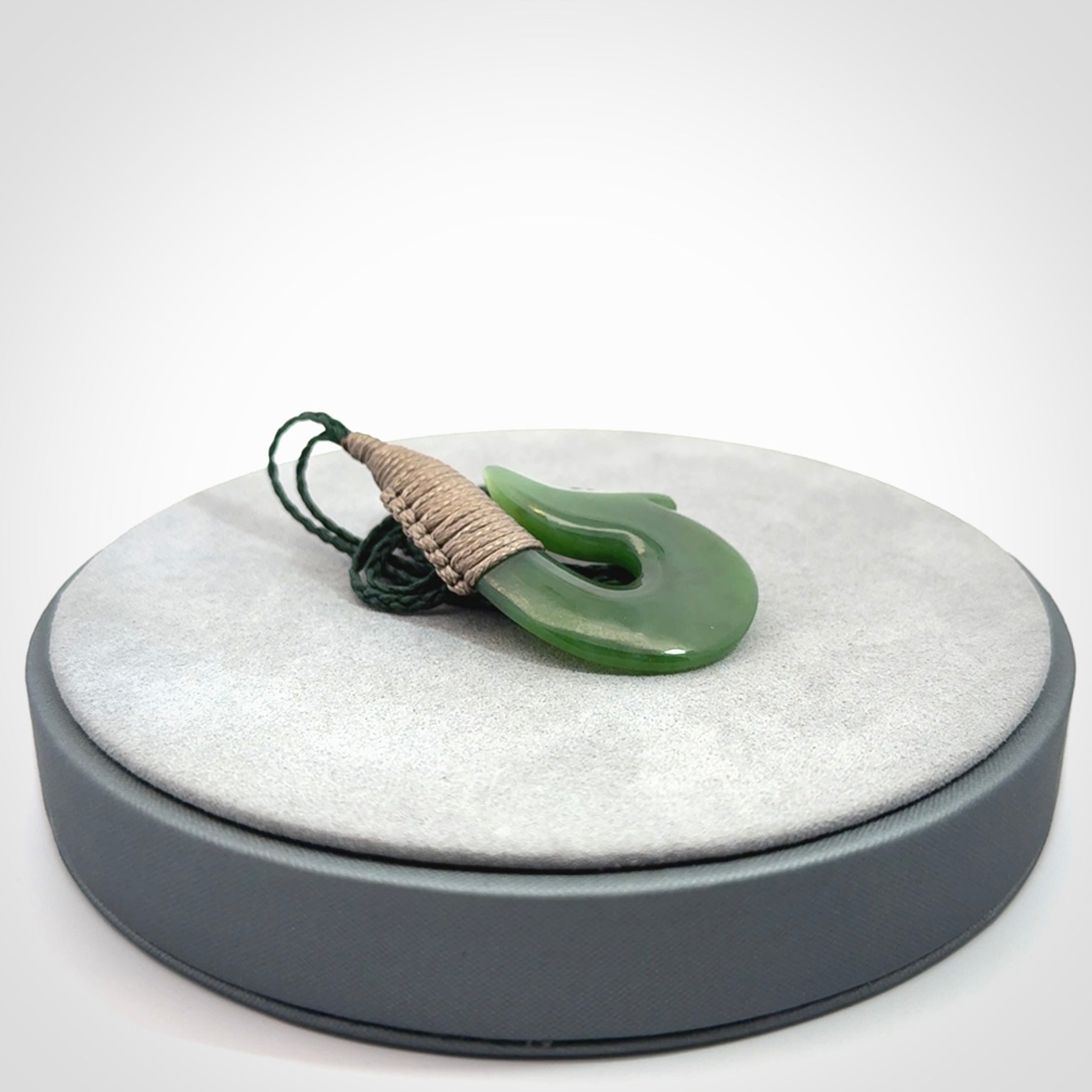 This photo shows a greenstone hook, or matau, pendant. It is a beautiful green British Columbia jade stone. The plaited cord is a green colour. It is plaited and the length can be adjusted.