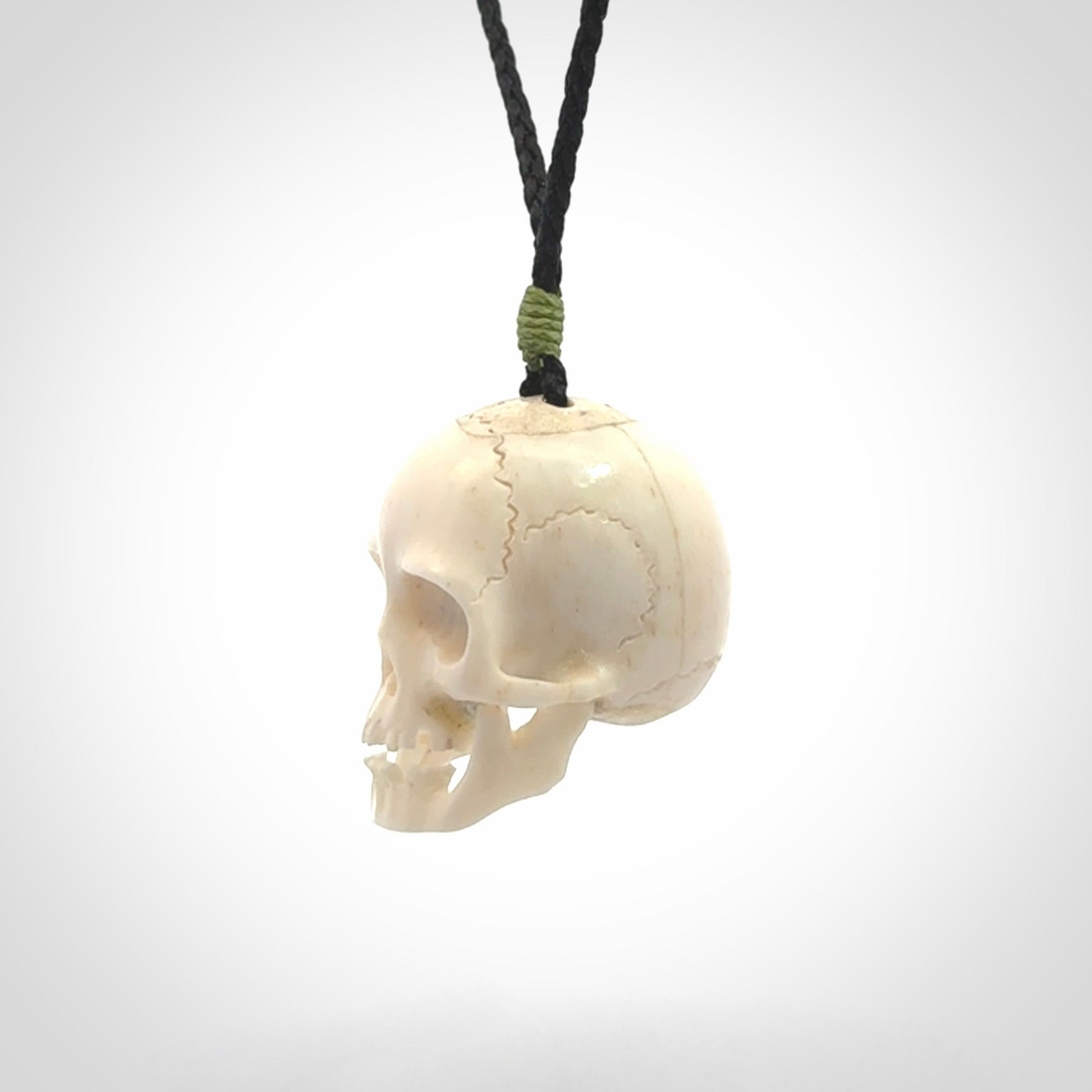 This picture shows a large sized Deer Antler Bone skull pendant that we have hand carved. It is polished to a soft shine and is a very striking piece. The cord is black and has a small green popper binding just above the pendant.