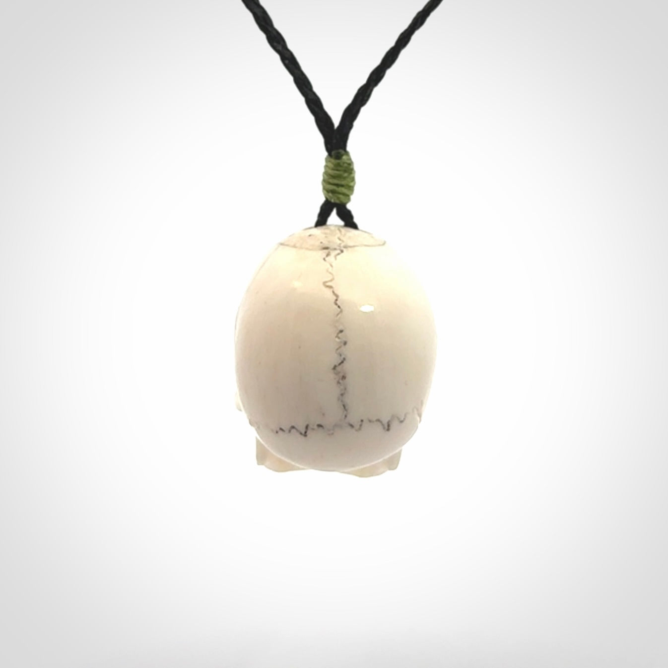 This picture shows a large sized Deer Antler Bone skull pendant that we have hand carved. It is polished to a soft shine and is a very striking piece. The cord is black and has a small green popper binding just above the pendant.