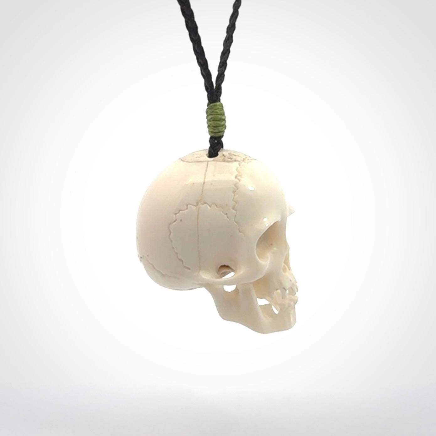 This picture shows a large sized Deer Antler Bone skull pendant that we have hand carved. It is polished to a soft shine and is a very striking piece. The cord is black and has a small green popper binding just above the pendant.