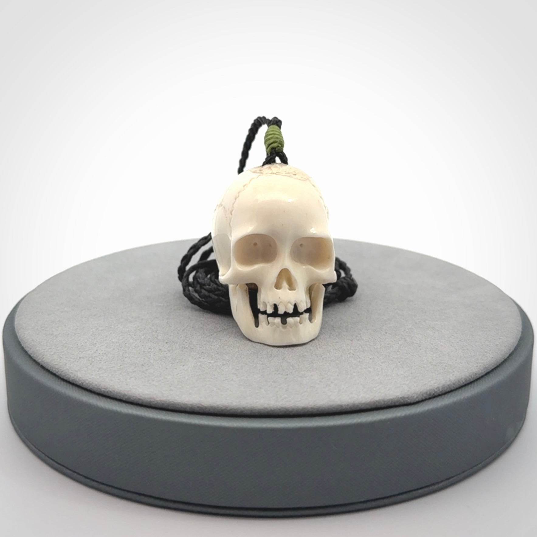 This picture shows a large sized Deer Antler Bone skull pendant that we have hand carved. It is polished to a soft shine and is a very striking piece. The cord is black and has a small green popper binding just above the pendant.