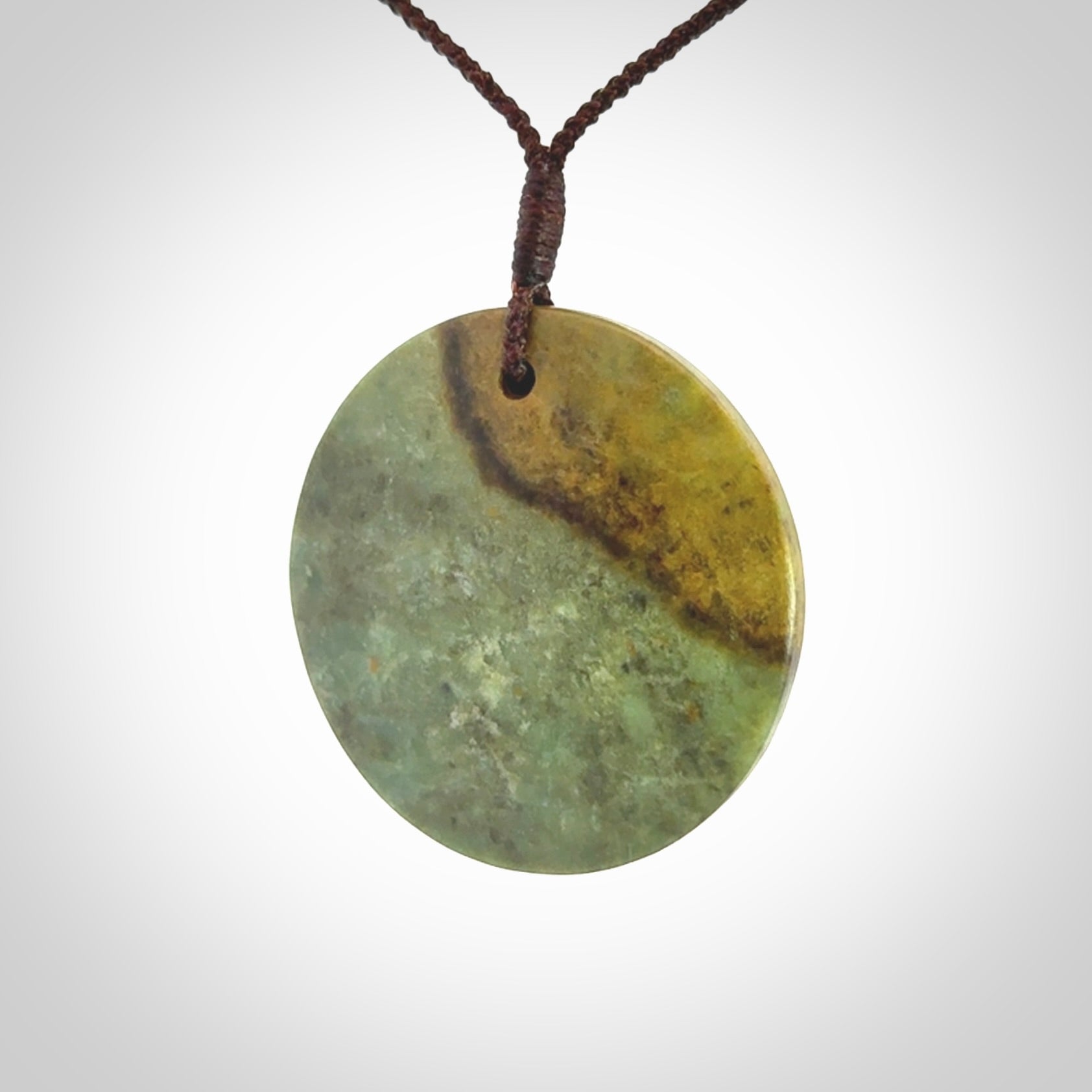 This piece is a large sized, oval round, disc pendant. It is suspended on a chocolate brown coloured braided cord that is length adjustable.