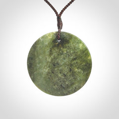 This piece is a large sized, oval round, disc pendant. It is suspended on a brown coloured braided cord that is length adjustable.