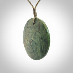 This piece is a large sized, oval round, disc pendant. It is suspended on a khaki coloured braided cord that is length adjustable.