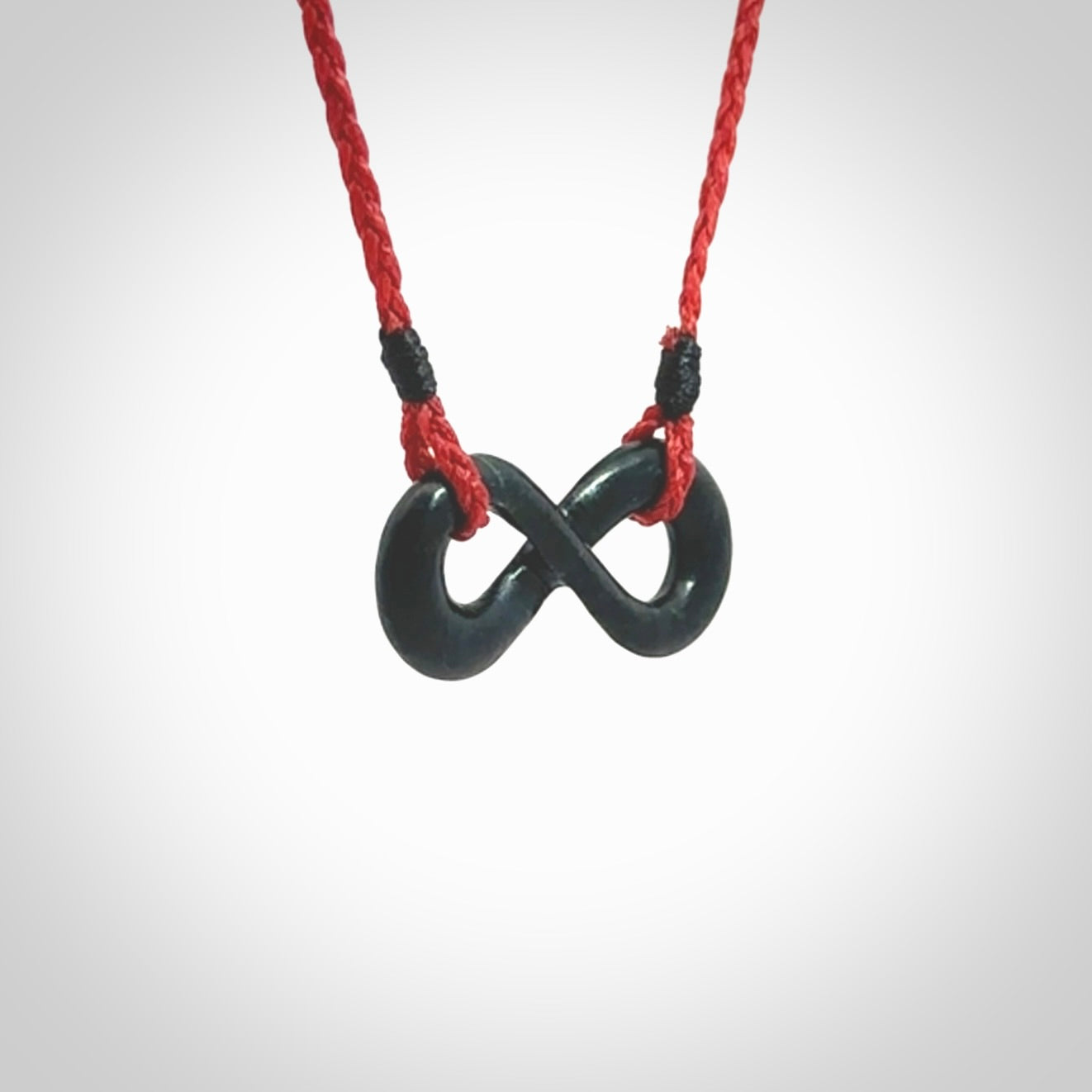 This piece is a fine, delicate eternity shaped pendant. It was carved from a lovely dark piece of black jade. It is suspended on an red braided cord that is length adjustable.