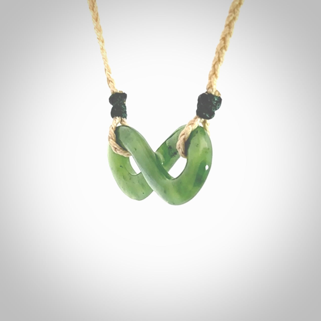 This piece is a fine, delicate eternity shaped pendant. It was carved from a lovely green piece of British Columbian jade. It is suspended on an oat coloured cord that is length adjustable.