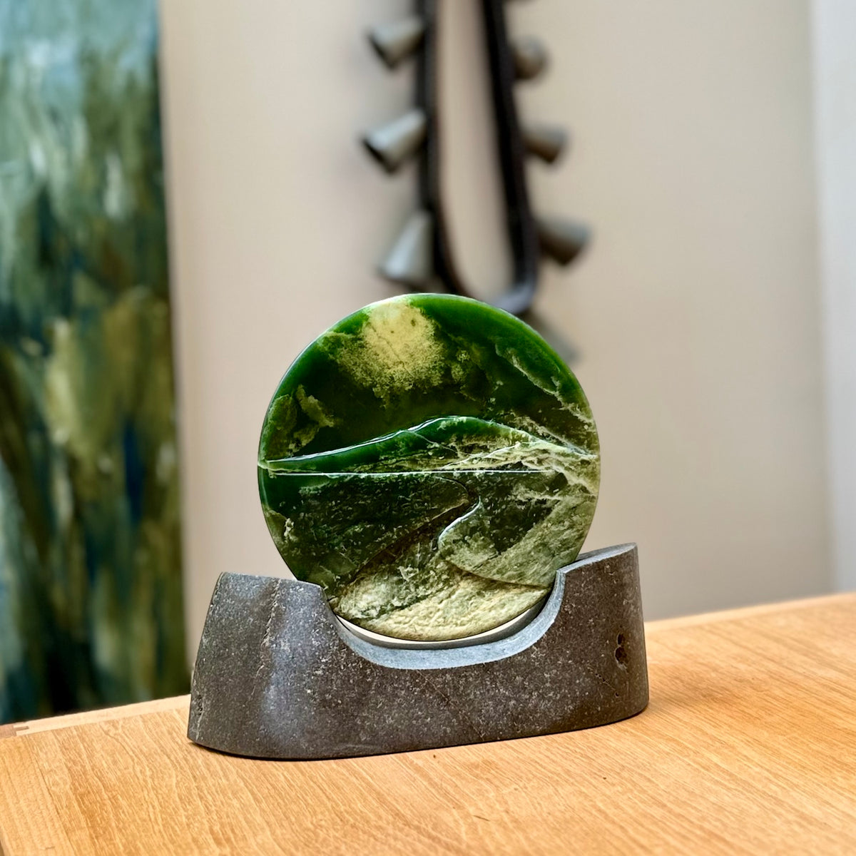 A spectacular, engraved jade disc sculpture. Hand carved in New Zealand from stunning Jade. This is a beautiful disc sculpture carved with a mountain and valley into its face. It is presented with a greywacke beach stone display stand. A beautiful sculptural piece hand carved in New Zealand.