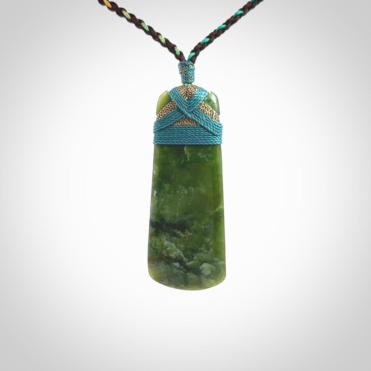 This photo shows a beautiful New Zealand jade toki suspended from a hand plaited adjustable cord. The binding around the top of the toki is a blue and bronze colour. The cord is length adjustable which allows the pendant to be positioned where it suits you best. The toki was carved for us by Jen Hung who is a wonderful jade carver from New Zealand.