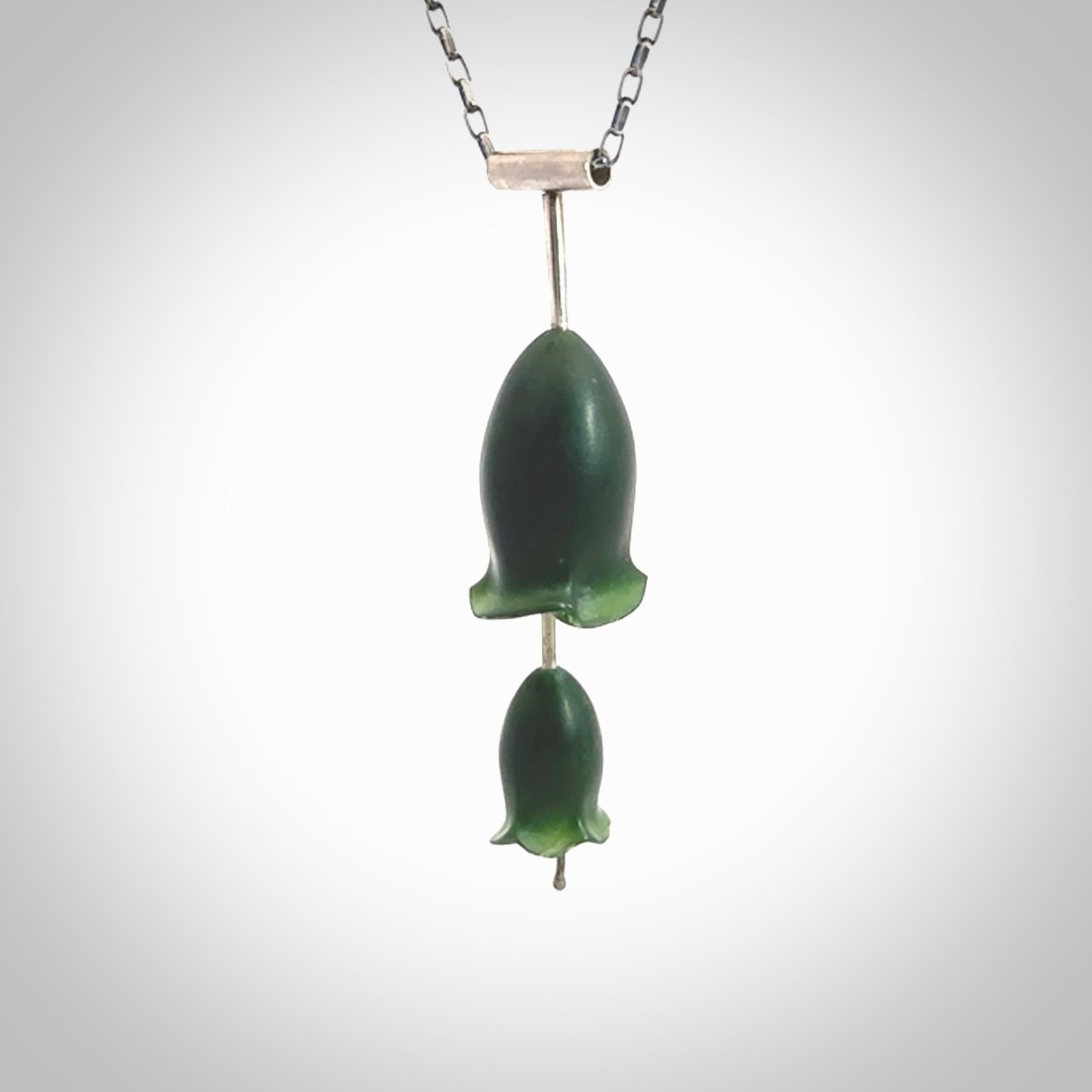This is a very dark green flower bud pendant carved here in New Zealand by Josey Coyle. It is handmade carved jade double flower buds one placed above the other on a fine silver wire. The top is a T-shaped tube through which a silver necklace chain passes. The jade florets are handcarved from beautiful dark green New Zealand Marsden Jade and the chain is handmade from sterling silver wire.