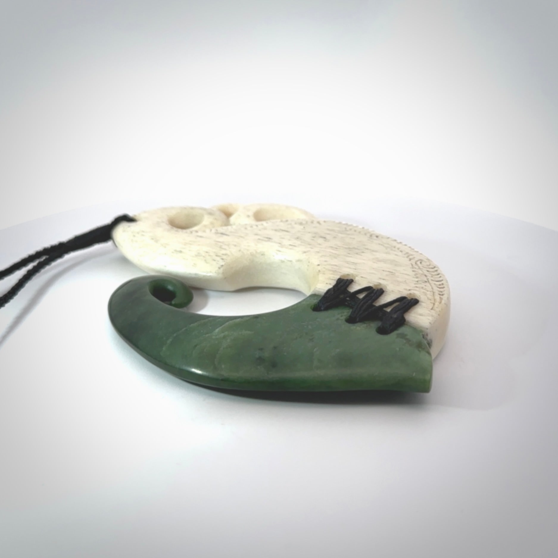 This photo shows a wonderful hand carved New Zealand jade and Whale Bone Manaia with hook pendant. The hook is suspended on a black four plait cord that is length adjustable.