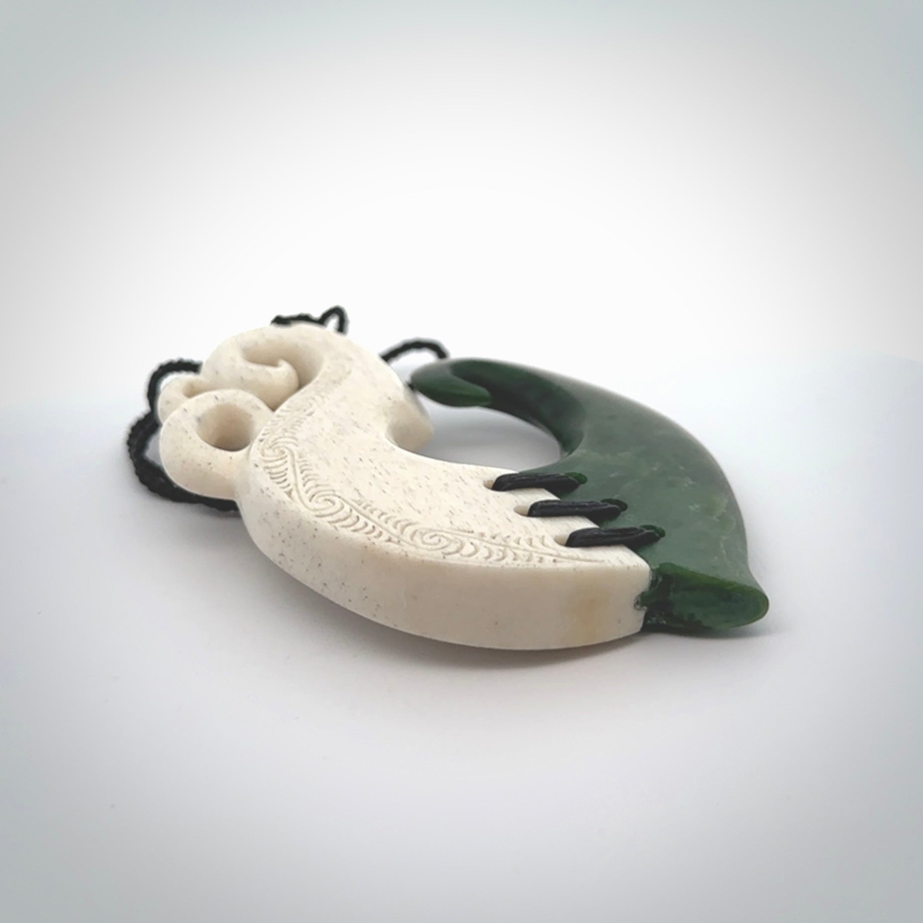 This photo shows a wonderful hand carved New Zealand jade and Whale Bone Manaia with hook pendant. The hook is suspended on a black four plait cord that is length adjustable.
