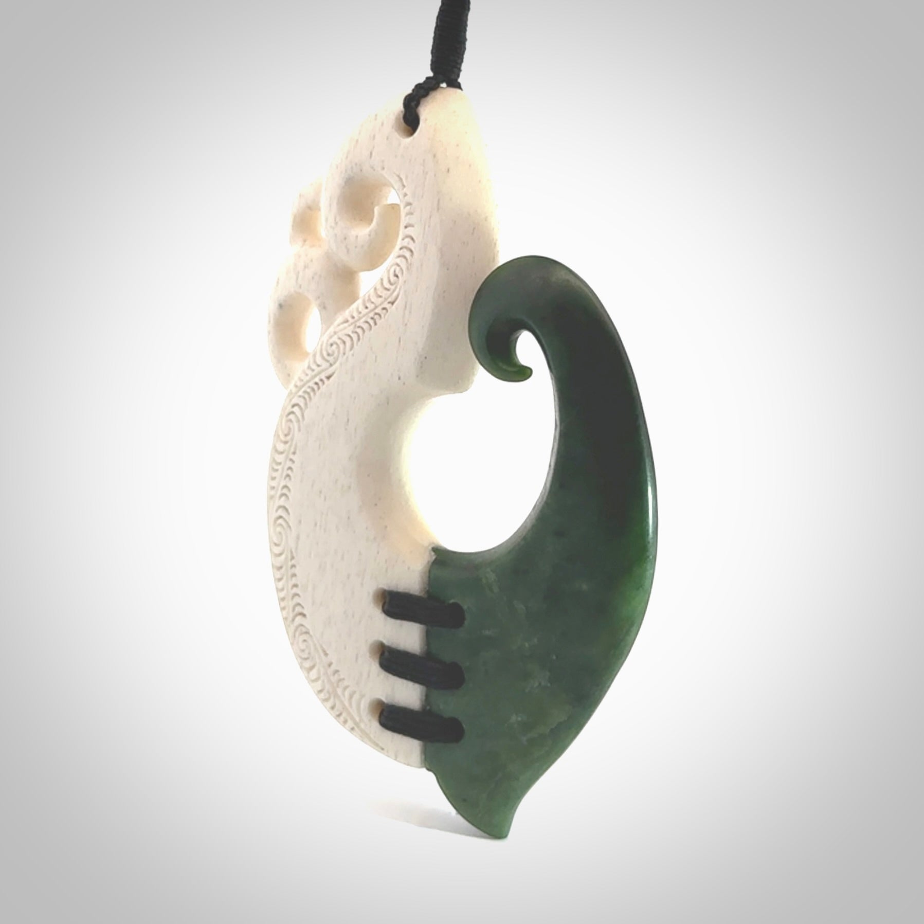 This photo shows a wonderful hand carved New Zealand jade and Whale Bone Manaia with hook pendant. The hook is suspended on a black four plait cord that is length adjustable.