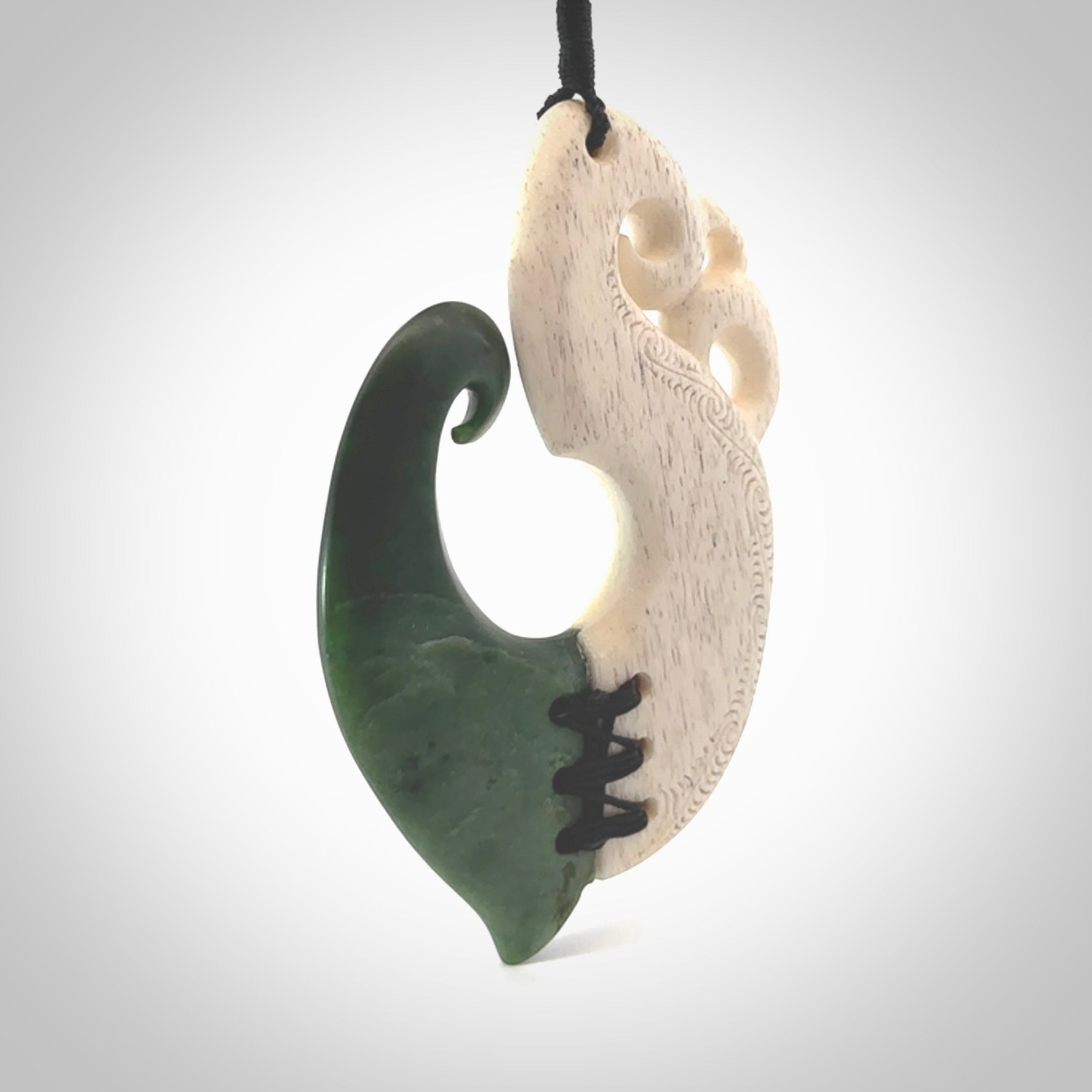 This photo shows a wonderful hand carved New Zealand jade and Whale Bone Manaia with hook pendant. The hook is suspended on a black four plait cord that is length adjustable.