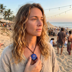 Woman on a beach holding a blue lapis lazuli pendant, with a crowd and ocean in the background
