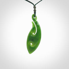 This photo shows a large matau, or hook, pendant carved in deep green New Zealand Jade. It is a very interesting piece which has a koru carved into the centre of the hook. It is strung on a manuka green cord which is adjustable. The artist who carved this pendant is Ross Crump, a master carver of jade from New Zealand.