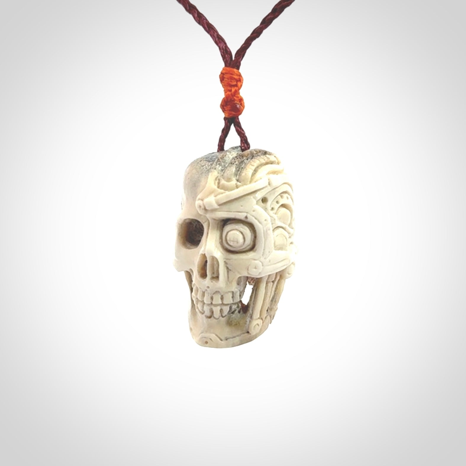 This picture shows a medium sized Deer Antler Bone robot skull pendant that we have hand carved. The cord is a plum colour and has a small orange binding just above the pendant.