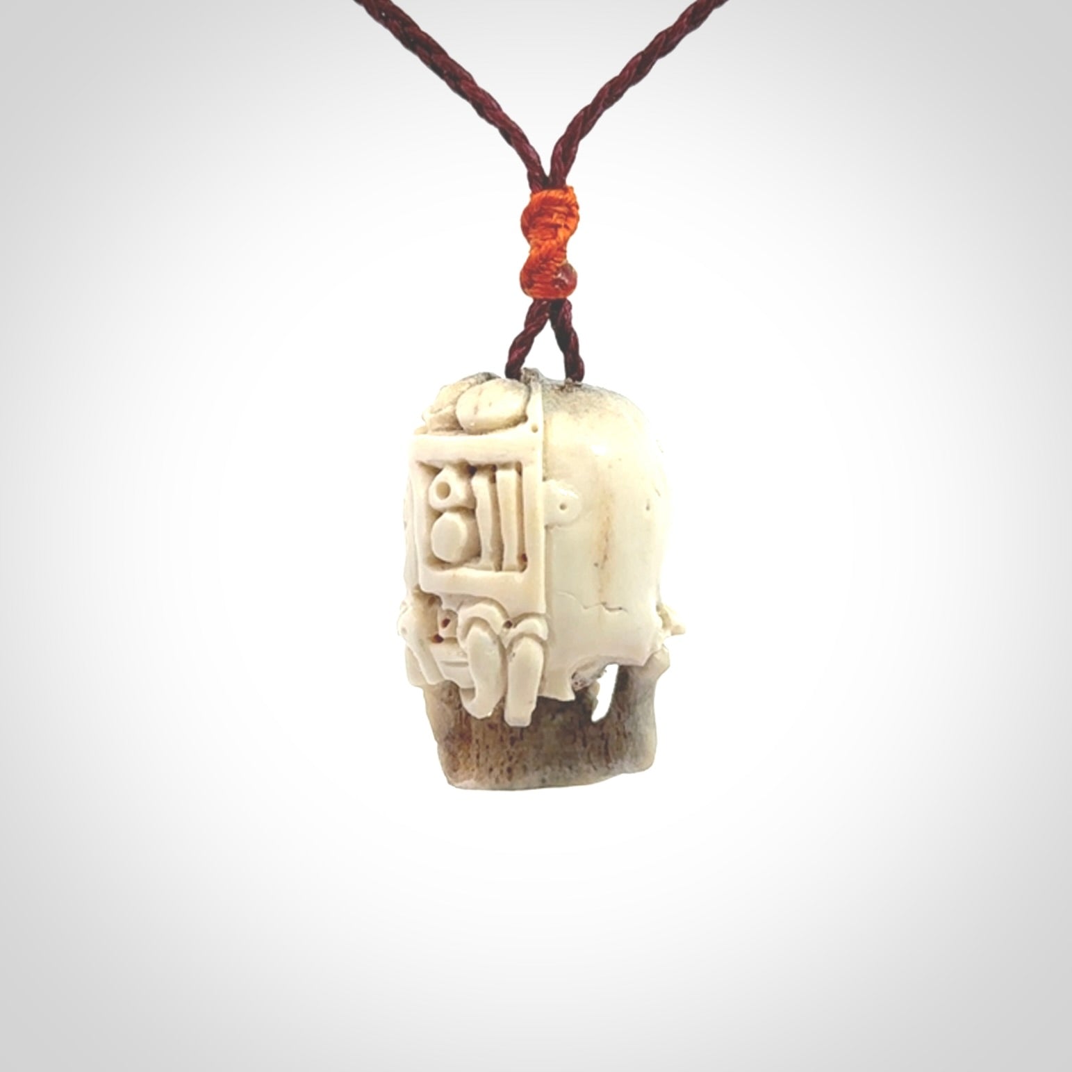This picture shows a medium sized Deer Antler Bone robot skull pendant that we have hand carved. The cord is a plum colour and has a small orange binding just above the pendant.