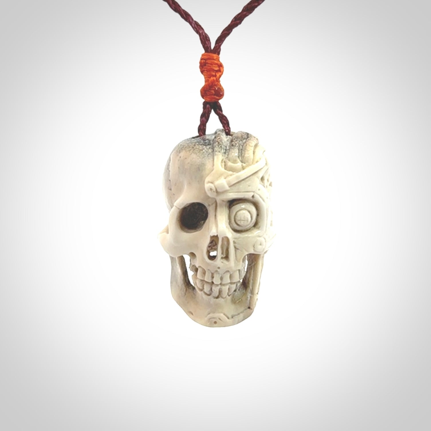 This picture shows a medium sized Deer Antler Bone robot skull pendant that we have hand carved. The cord is a plum colour and has a small orange binding just above the pendant.