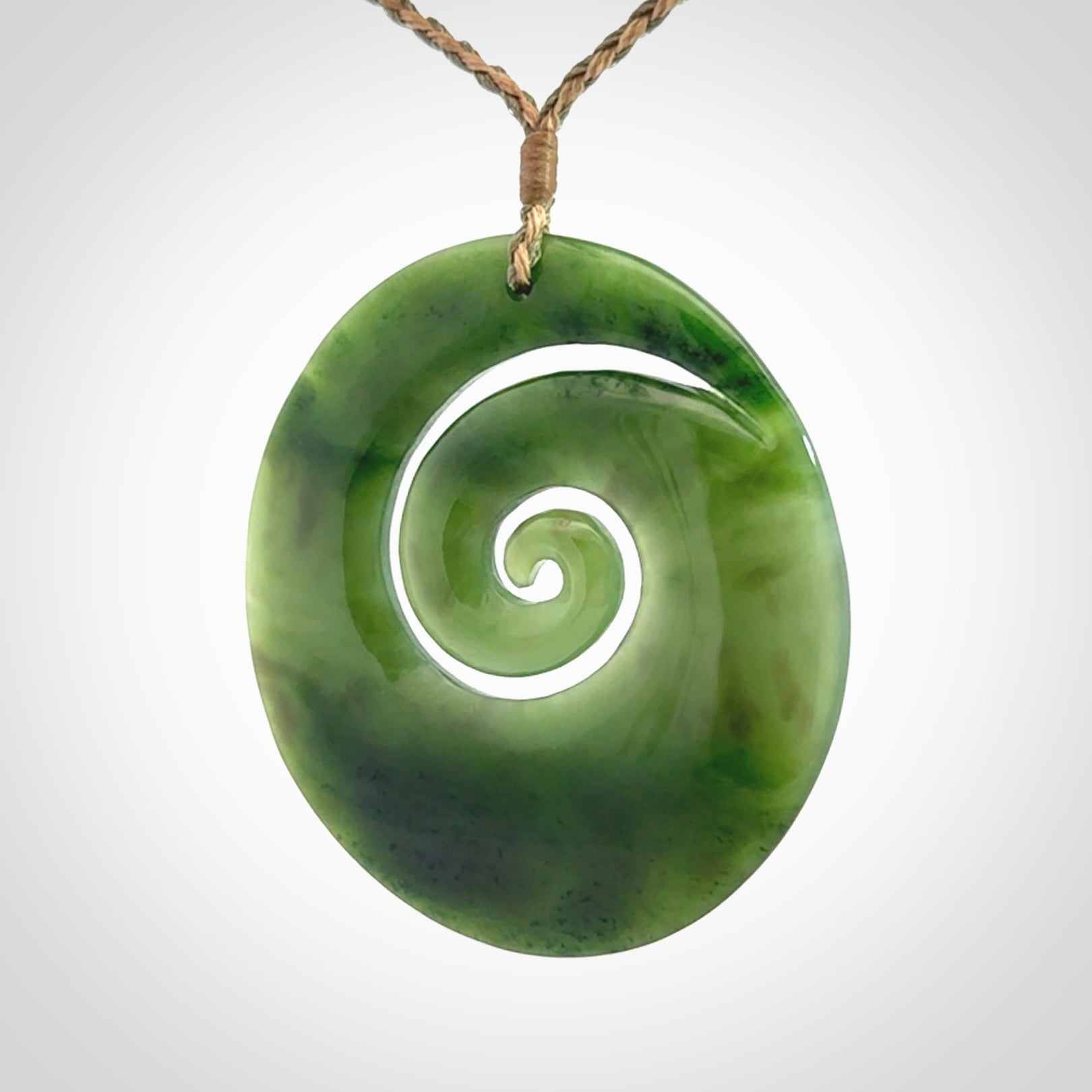 This picture shows a large hand-carved koru. It is carved from British Columbian jade and is suspended from a hand-plaited, tan/green cord.