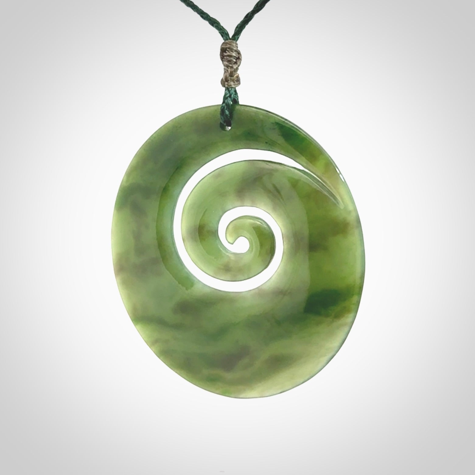 This picture shows a large hand-carved koru. It is carved from British Columbian jade and is suspended from a hand-plaited, green cord.