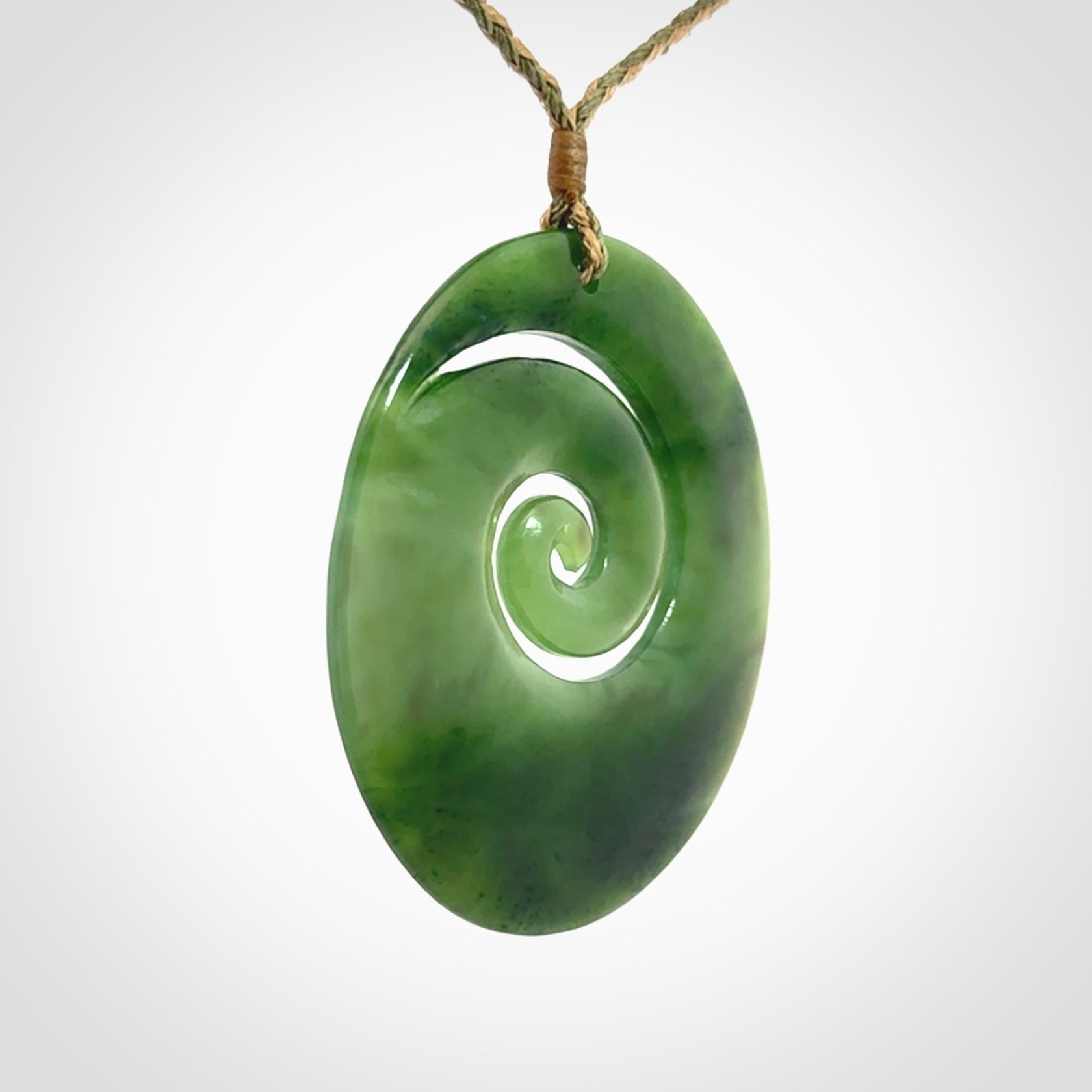 This picture shows a large hand-carved koru. It is carved from British Columbian jade and is suspended from a hand-plaited, tan/green cord.