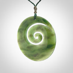 This picture shows a large hand-carved koru. It is carved from British Columbian jade and is suspended from a hand-plaited, green cord.