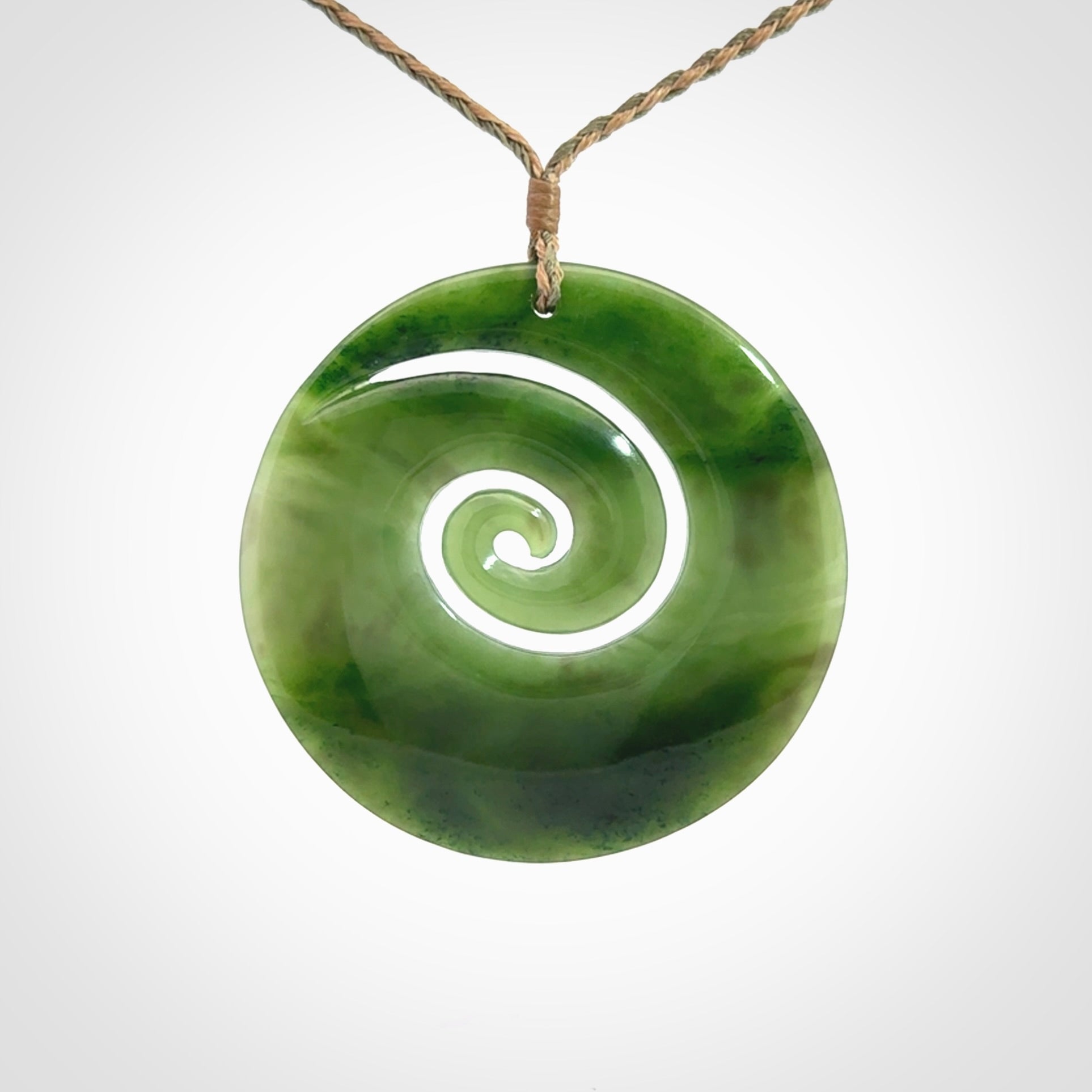 This picture shows a large hand-carved koru. It is carved from British Columbian jade and is suspended from a hand-plaited, tan/green cord.