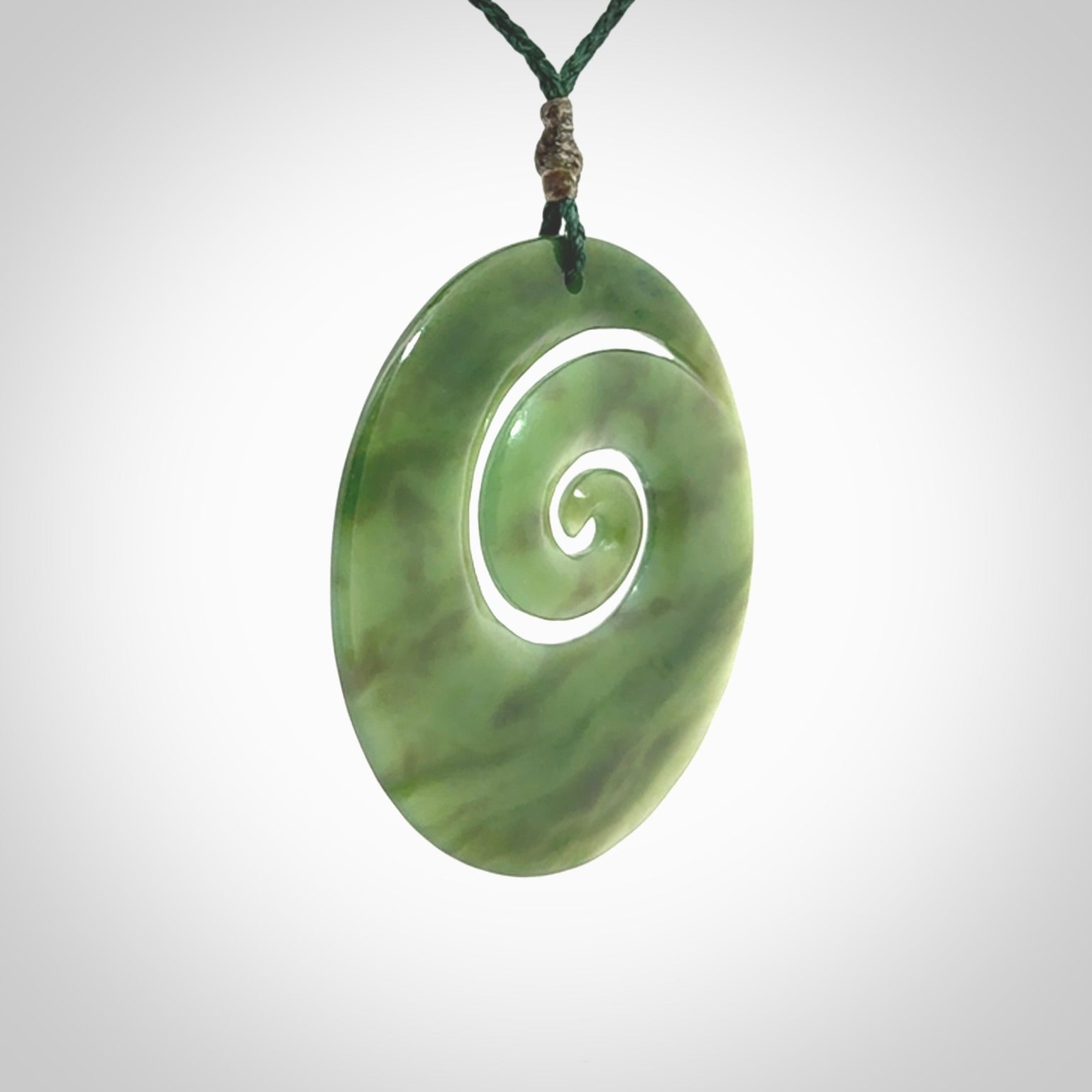 This picture shows a large hand-carved koru. It is carved from British Columbian jade and is suspended from a hand-plaited, green cord.