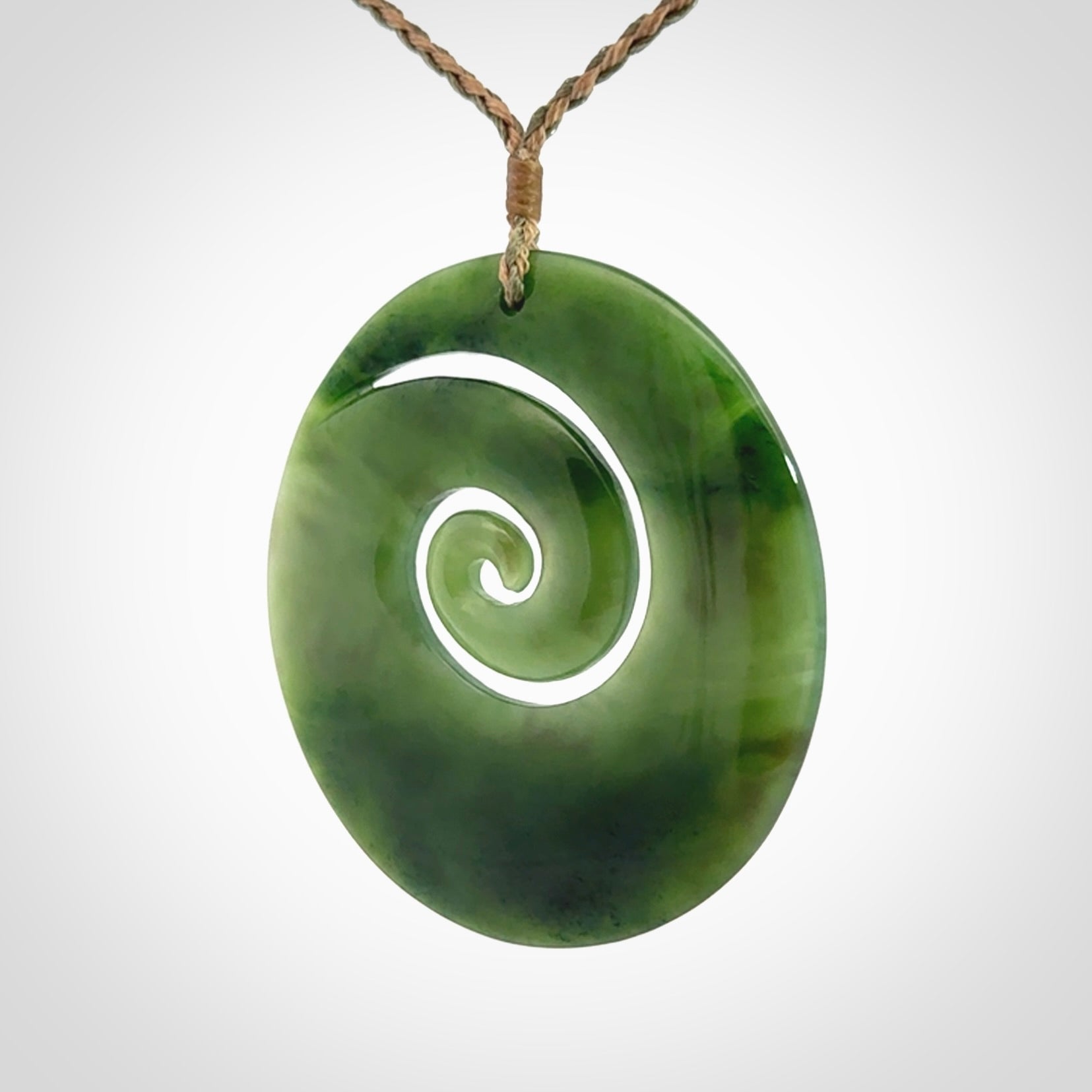This picture shows a large hand-carved koru. It is carved from British Columbian jade and is suspended from a hand-plaited, tan/green cord.